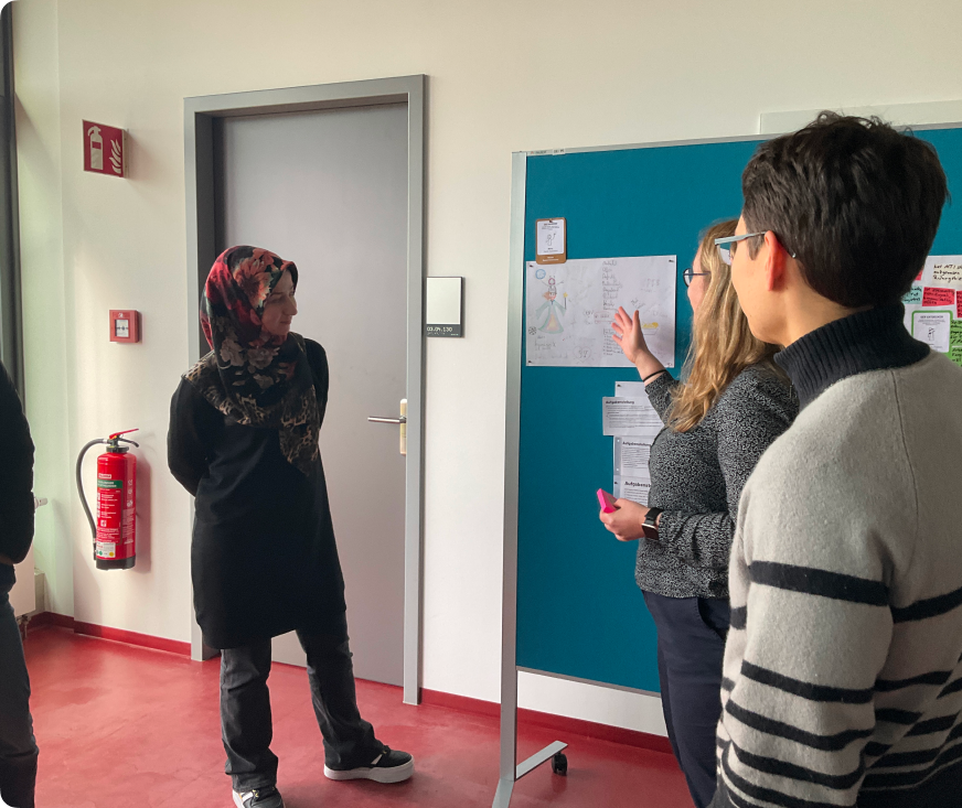 Das Bild zeigt eine Gruppe von vier Personen, die in einem Besprechungsraum stehen. Eine Frau mit einem Hijab in Rot- und Schwarztönen steht links und schaut auf ein Whiteboard. Eine zweite Frau mit langen, blonden Haaren und Brille steht neben dem Whiteboard und erklärt etwas, während sie auf eine Zeichnung zeigt. Rechts von ihr steht ein Mann mit dunklen Haaren und Brille, der einen grauen Pullover mit schwarzen Streifen trägt. Eine vierte Person, teilweise im Bild, steht links und hört zu. Im Hintergrund sind eine geschlossene graue Tür, ein Feuerlöscher und ein Fenster zu sehen. Das Whiteboard ist mit verschiedenen Zeichnungen und Notizen versehen.