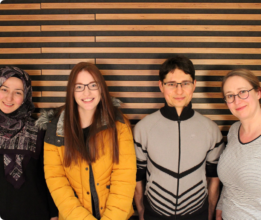 Das Bild zeigt das Team von MTE. Es sind vier Personen zu sehen, die vor einer Holzwand mit horizontalen Streifen stehen. Von links nach rechts sind das:

Eine Frau mit einem Hijab in verschiedenen Lila- und Grautönen, die lächelt.
Eine Frau mit langen, glatten, roten Haaren und einer Brille, die ebenfalls lächelt und eine gelbe Winterjacke trägt.
Ein Mann mit kurzen, dunklen Haaren und einer Brille, der einen grauen Pullover mit schwarzen Streifen trägt und lächelt.
Eine Frau mit blonden, zurückgebundenen Haaren und einer Brille, die ein gestreiftes Oberteil trägt und ebenfalls lächelt.
Alle vier Personen stehen nebeneinander und blicken in die Kamera.